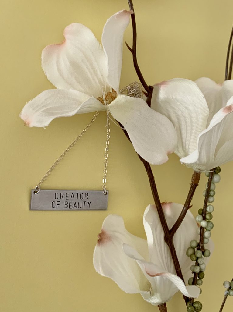 A necklace engraved with "creator of beauty" hanging from a flowering plant