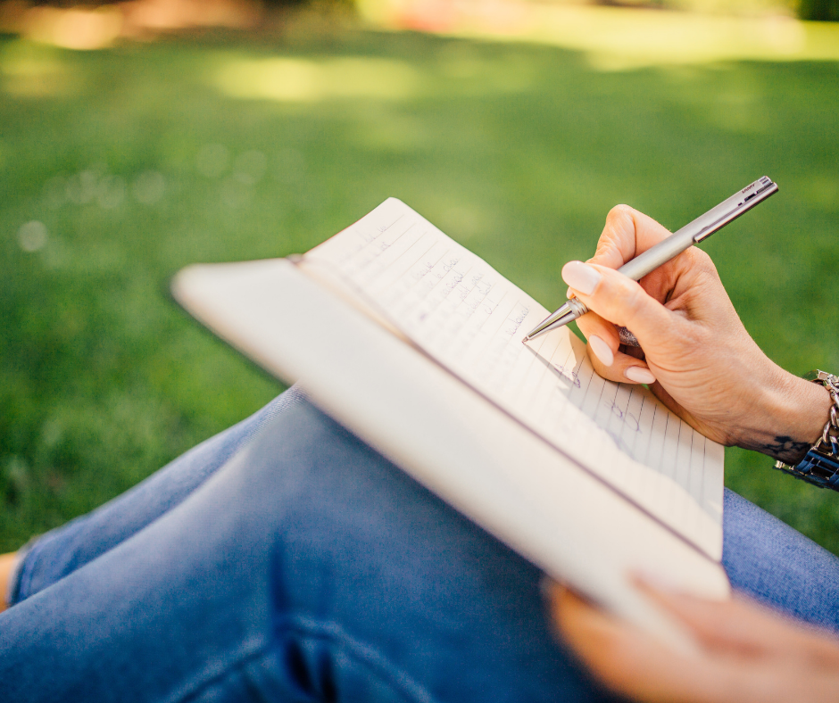 Tampa's Copywriter sitting in a grass field and writing in a notebook for "Why Me?' blog post.